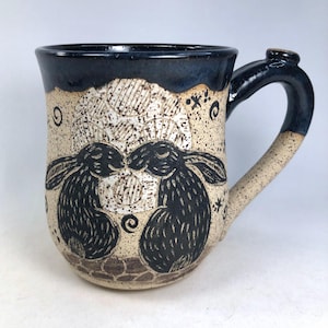 May include: A ceramic mug with a dark blue rim and handle. The mug features a speckled beige background with a black illustration of two kissing rabbits. The handle has a small thumb rest.