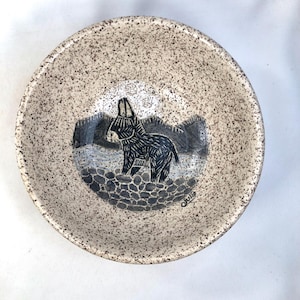 May include: A speckled ceramic bowl with a hand-drawn image of a donkey. The donkey is black and stands on a gray stone surface, with a gray and blue background. The bowl's rim is speckled with brown.