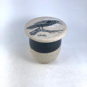 May include: A ceramic container with a lid featuring a black raven design perched on a branch. The container has a speckled beige finish with a black band around the middle. The lid has the name "julie" written on it.