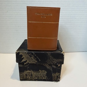Puede incluir: Una cartera de cuero marrón con el texto "Travellers Aid" en letras doradas. La cartera tiene dos rayas horizontales doradas y está sobre una caja negra con un diseño arquitectónico dorado.