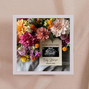 May include: A white card with the text "We're Expecting!" and a black and white ultrasound image. The card also says "Baby Stevenson" and "February 2023". The card is surrounded by a bouquet of pink, yellow, and orange flowers.