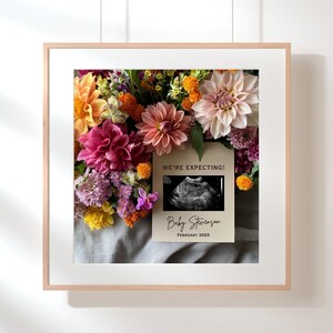 May include: A framed print featuring a floral arrangement of pink, orange, and yellow flowers. A card with the text "We're Expecting!" and an ultrasound image is placed on top of the flowers. The card also includes the text "Baby Stevenson, February 2023."