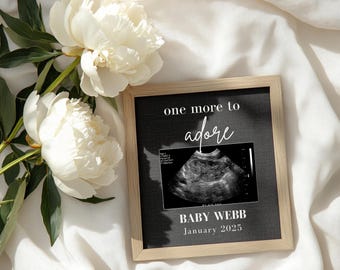 Puede incluir: Una imagen de ultrasonido en blanco y negro en un marco de madera con el texto "one more to adore" y "BABY WEBB January 2025".