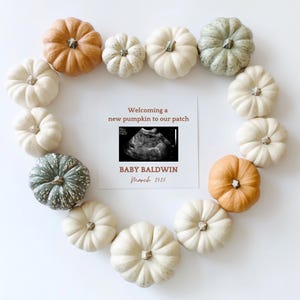 Pode incluir: Um cartão branco com o texto "Welcoming a new pumpkin to our patch" e uma imagem de ultrassom em preto e branco. O cartão também diz "Baby Baldwin March 2025". O cartão é cercado por um formato de coração feito de pequenas abóboras brancas e laranja.