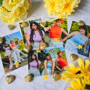 May include: A collection of photographs featuring women in various outfits, including crop tops and skirts, arranged on a white surface. The photos are adorned with gold glitter hearts, star-shaped lights, and yellow flowers, creating a vibrant and artistic display.