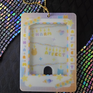 May include: A rectangular, clear plastic charm holder with a gold chain. The holder features pastel colors, including yellow, blue, and purple, with the words "K Charm Affect" printed on it. The design includes stars, hearts, and bear illustrations.