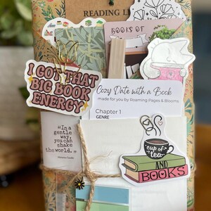 May include: A wrapped book gift with decorative stickers and a gold paperclip. Stickers include text that says "I GOT THAT BIG BOOK ENERGY", "cup of tea AND BOOKS", and "Cozy Date with a Book". The gift is tied with twine.
