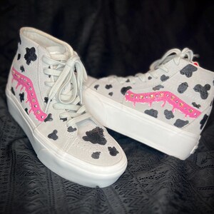 May include: White high-top sneakers with a black and white cow print design. The sneakers have pink dripping paint with rhinestones on the side.