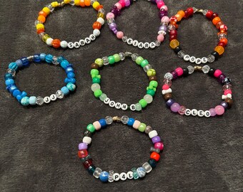 Puede incluir: Un conjunto de 8 pulseras de cuentas coloridas con diferentes nombres escritos en ellas. Las pulseras están hechas con cuentas de plástico de colores y cuentas transparentes. Los nombres en las pulseras son Sarah, Kiara, Pope, JJ, John B, Cleo y P4L.