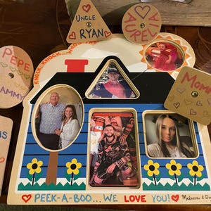 Può includere: Una cornice per foto in legno a forma di casa con più aperture per foto. La cornice è dipinta di blu e ha un motivo di fiori gialli. La cornice ha un cartello che dice "Peek-a-boo... We love you! Melissa & Doug".