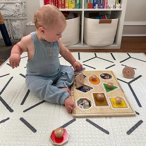 May include: A wooden shape puzzle with various shapes and knobs, featuring family photos. The puzzle is on a patterned play mat. A baby in a blue jumpsuit is interacting with the puzzle. A wooden toy with the word "Mommy" is also visible.