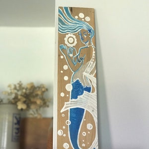 Puede incluir: Un letrero de madera con una sirena pintada a mano con cabello azul y cola. La sirena sostiene una concha y est&aacute; rodeada de burbujas blancas.