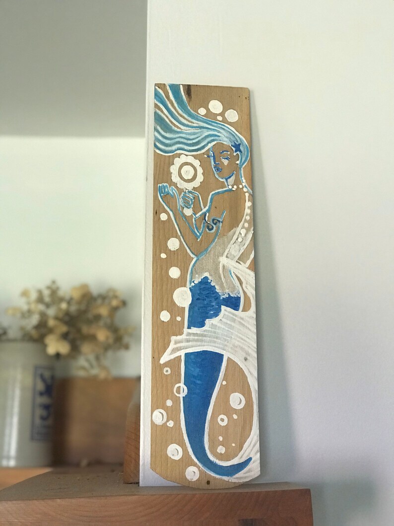Puede incluir: Un letrero de madera con una sirena pintada a mano con cabello azul y cola. La sirena sostiene una concha y est&aacute; rodeada de burbujas blancas.