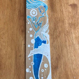 Puede incluir: Una sirena azul y blanca pintada en una tabla de madera. La sirena tiene el pelo largo y suelto y sostiene una perla en su mano. Hay burbujas blancas a su alrededor.