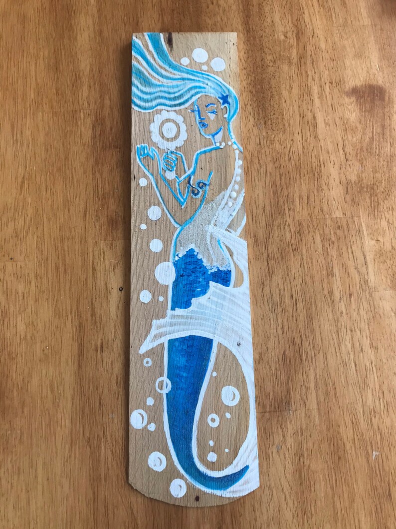 Puede incluir: Una sirena azul y blanca pintada en una tabla de madera. La sirena tiene el pelo largo y suelto y sostiene una perla en su mano. Hay burbujas blancas a su alrededor.