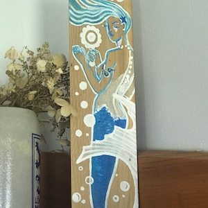 Puede incluir: Una sirena azul y blanca pintada en una tabla de madera. La sirena tiene el pelo largo y fluido y sostiene una flor blanca. El fondo es de madera clara con burbujas blancas.
