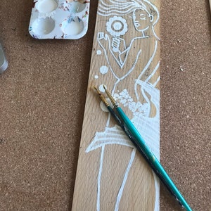 Puede incluir: Una tabla de madera con un dise&ntilde;o pintado en blanco de una mujer con el pelo suelto sosteniendo una flor. La mujer est&aacute; pintada en un estilo caprichoso con un contorno simple. Dos pinceles est&aacute;n sobre la tabla.