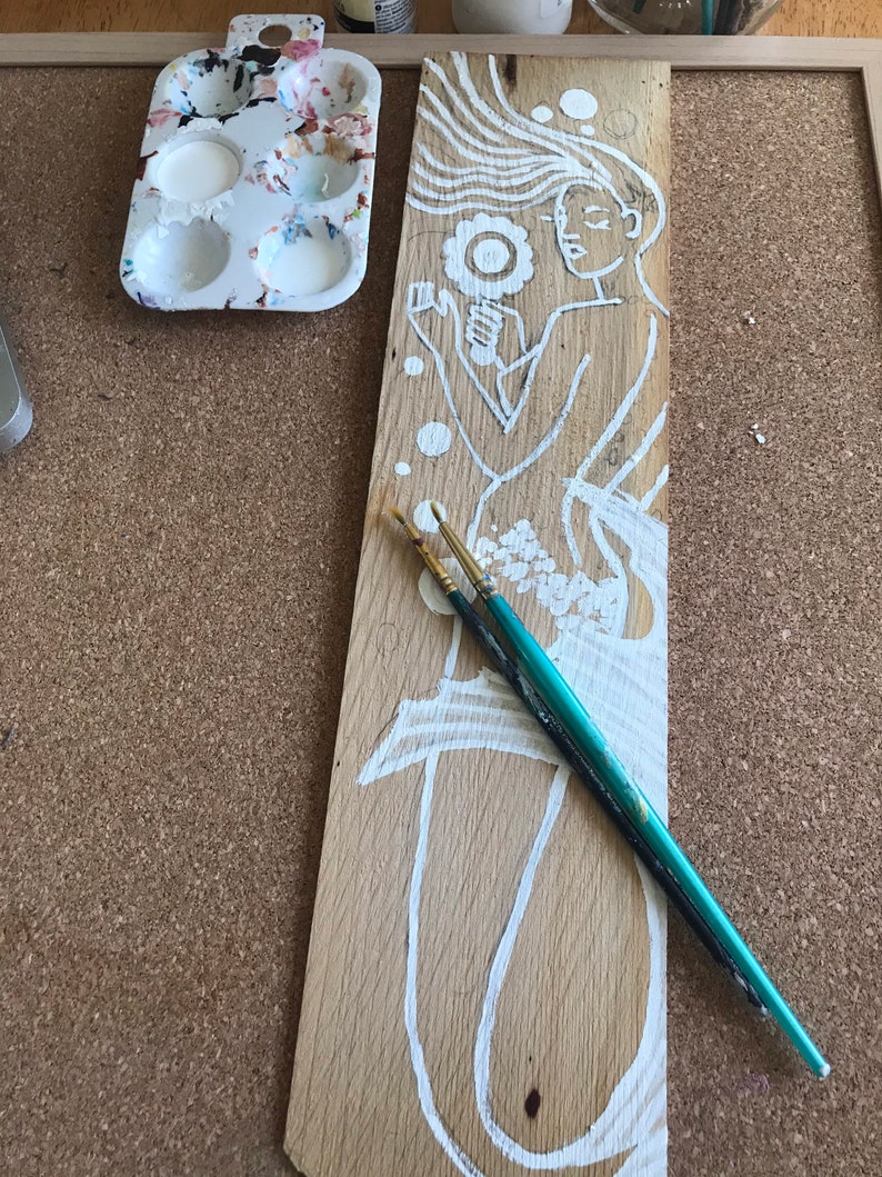 Puede incluir: Una tabla de madera con un dise&ntilde;o pintado en blanco de una mujer con el pelo suelto sosteniendo una flor. La mujer est&aacute; pintada en un estilo caprichoso con un contorno simple. Dos pinceles est&aacute;n sobre la tabla.