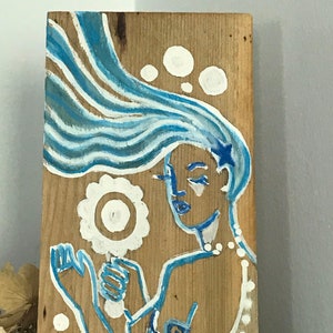 Puede incluir: Una pintura azul y blanca sobre una tabla de madera que representa una sirena con el pelo largo y suelto, sosteniendo una flor blanca. La sirena lleva un collar blanco y tiene una estrella azul en la cabeza.