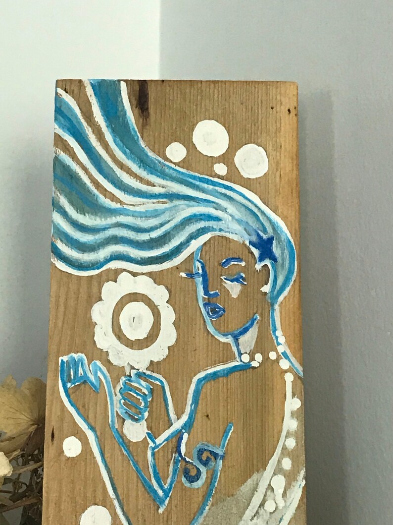 Puede incluir: Una pintura azul y blanca sobre una tabla de madera que representa una sirena con el pelo largo y suelto, sosteniendo una flor blanca. La sirena lleva un collar blanco y tiene una estrella azul en la cabeza.