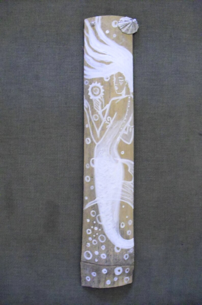 Puede incluir: Sirena pintada de blanco con cabello suelto y una concha en un fondo de tabl&oacute;n de madera. La sirena sostiene una flor blanca y est&aacute; rodeada de burbujas pintadas de blanco.