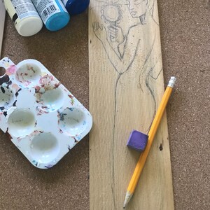 Puede incluir: Una tabla de madera con un dibujo a l&aacute;piz de una mujer sosteniendo un espejo. La tabla est&aacute; rodeada de materiales de arte, incluyendo pintura, una paleta de pintura y una goma de borrar.