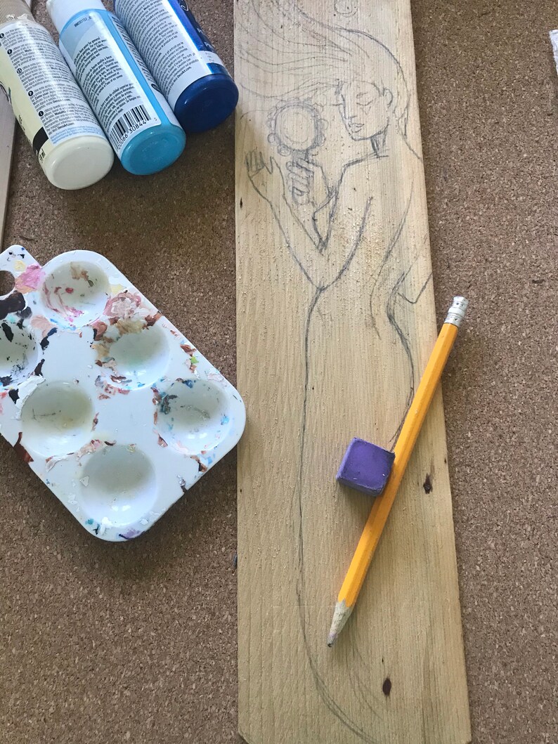 Puede incluir: Una tabla de madera con un dibujo a l&aacute;piz de una mujer sosteniendo un espejo. La tabla est&aacute; rodeada de materiales de arte, incluyendo pintura, una paleta de pintura y una goma de borrar.