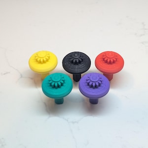 以下が含まれることがあります： 黄色、黒、赤、ティール、紫の5色のカラフルな装飾ノブ。各ノブは、詳細なデザインの円形の上部と円筒形のステムを備えています。ノブは白い表面に配置されています。