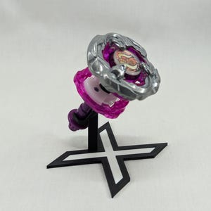 Può includere: Una trottola viola e argento su un supporto nero a forma di croce. La parte superiore presenta un design circolare e un anello esterno argentato. Il supporto è nero, su sfondo bianco.