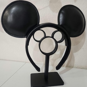 May include: Black mouse ear headband with a matching stand. The ears are round and padded, and the headband features a Mickey Mouse silhouette design. The stand is black and has a square base.