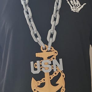 May include: A large, gray chain necklace with a gold anchor pendant and the letters "USN". The necklace is worn over a black t-shirt with a white skeleton hand graphic.