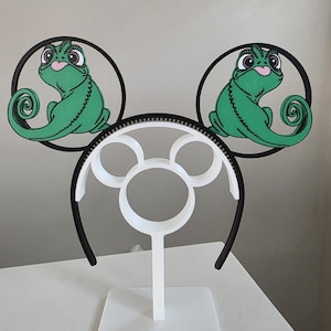 May include: A pair of black and white mouse ear headbands with green chameleon designs. The ears are circular with a black outline, and the chameleon is green with a pink tongue. The headband is supported by a white stand.