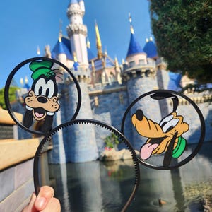 May include: Black headband featuring two circular cutouts with cartoon character designs. One cutout shows Goofy in a green hat, and the other shows Pluto with his tongue out. The background includes a castle and water.