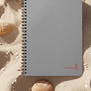 May include: A grey spiral-bound notebook with a red logo of a person paddleboarding. The notebook is on a sandy surface with seashells and small white pebbles. The notebook has a black spiral binding.