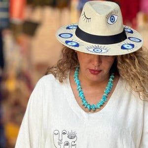 May include: A white straw hat with blue and black evil eye designs. The hat has a black band and a wide brim. The person wearing the hat has curly blonde hair and is wearing a white shirt with a turquoise necklace.