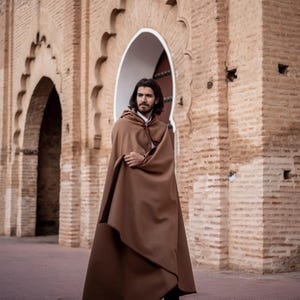 May include: A person wearing a brown hooded cloak, standing in front of a brick archway. The cloak is long and flowing, and the person is looking to the right.