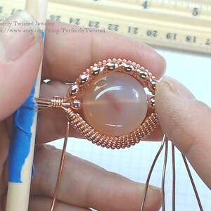May include: A close-up of a copper wire wrapped around a light pink agate gemstone. The wire is twisted and forms a decorative border around the stone. The stone is set on a wooden surface.