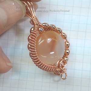 May include: A copper wire wrapped pendant featuring a round, light peach agate gemstone. The pendant has a copper wire loop for attaching to a chain or cord.