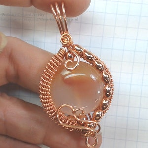May include: A copper wire wrapped pendant featuring a peach-colored agate gemstone. The pendant has a twisted wire design and a delicate swirl at the top.
