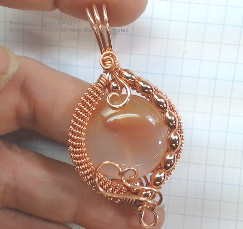May include: A copper wire wrapped pendant featuring a peach-colored agate gemstone. The pendant has a twisted wire design and a delicate swirl at the top.