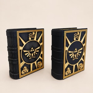 Peut inclure: Deux objets en forme de livre noir avec des emblèmes dorés en relief sur les couvertures avant. Chaque emblème présente un symbole Triforce central entouré de rayons solaires et de motifs circulaires, encadré par une bordure dorée.