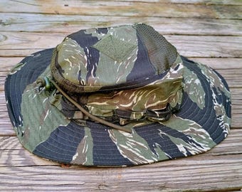 May include: A boonie hat featuring a tiger stripe camouflage pattern in shades of green, brown, and black. The hat includes a wide brim, a flat top with a square hook-and-loop patch, mesh side panels, and an adjustable chin cord with a toggle.