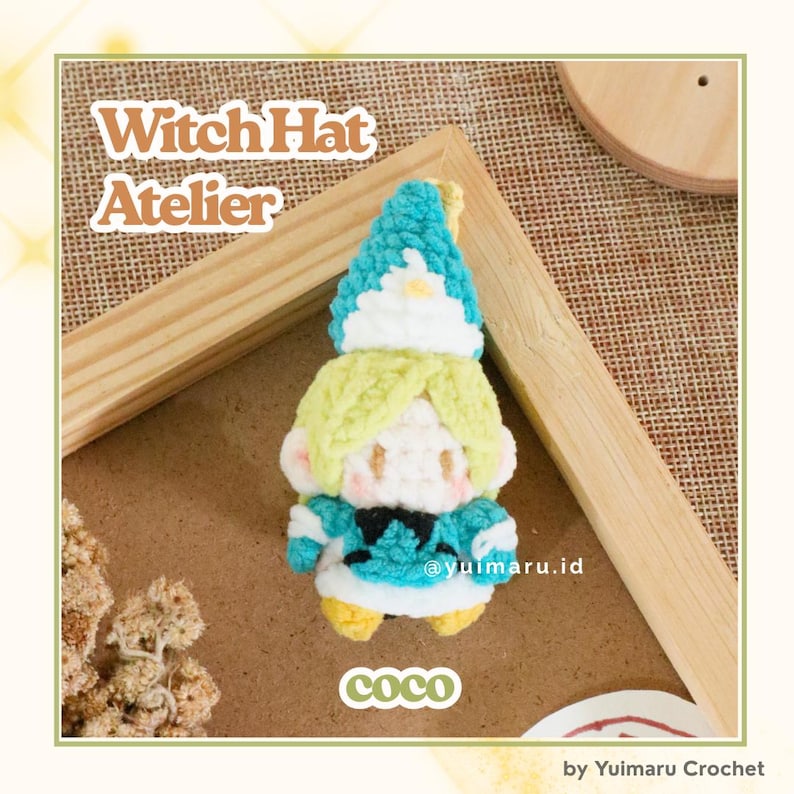 May include: A small, handmade crochet doll featuring a pointed teal hat with a white brim, light yellow hair, and a teal outfit with black accents. The doll has a white face with simple features and yellow feet. Text reads Witch Hat Atelier and coco.