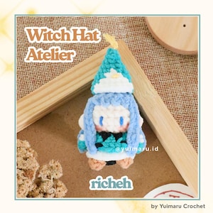 May include: A small crochet figure of a character wearing a pointed teal and white witch hat, light blue hair, and a teal patterned outfit. The text Witch Hat Atelier and richeh are visible. The figure is crafted with yarn in shades of teal, white, and tan.