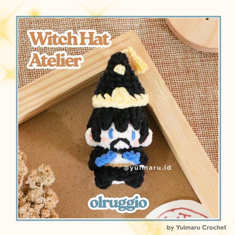 May include: A small, handmade crochet doll featuring a black witch hat with a yellow band and tassel, black hair, blue eyes, and a black outfit with blue accents. The text "Witch Hat Atelier" and "olruggio" are visible in the image.