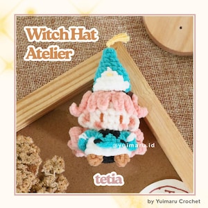 May include: A handmade crochet doll featuring a teal pointed witch hat with a white brim, light pink hair, and a teal and white outfit. The text Witch Hat Atelier and tetia are visible. The doll is crafted with soft yarn and intricate stitching.