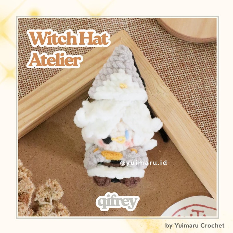 May include: A small, handmade crochet plush figure featuring a pointed gray hat, fluffy white hair, and a white and gray outfit with a yellow accent. The figure has a black eye patch and is labeled with the text Witch Hat Atelier and qifrey.