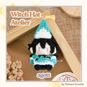 May include: A small, handmade crochet plush figure featuring a teal and white pointed hat, black hair, and a teal and white outfit. The figure has a light-colored face with subtle pink cheeks. Text on the image reads Witch Hat Atelier and agott.