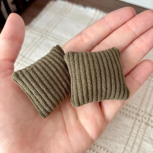 May include: Two miniature square pillows in a solid olive green color. Each pillow features a distinct vertical ribbed knit texture across the entire surface, creating a soft and uniform appearance for these small decorative items.