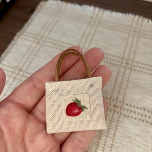 Op de afbeelding: Een vierkante miniatuur draagtas van gebroken wit, gestructureerd textiel met een centraal geborduurde rode aardbei met een groen blaadje. De tas heeft twee dunne, bruine, glinsterende hengsels bevestigd aan de bovenrand van de tas.